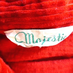 May include: A bright red corduroy garment featuring a white fabric label sewn into the collar area. The label displays the word Majestic embroidered in a cursive green font. The corduroy fabric has a distinct ribbed texture throughout the surface.
