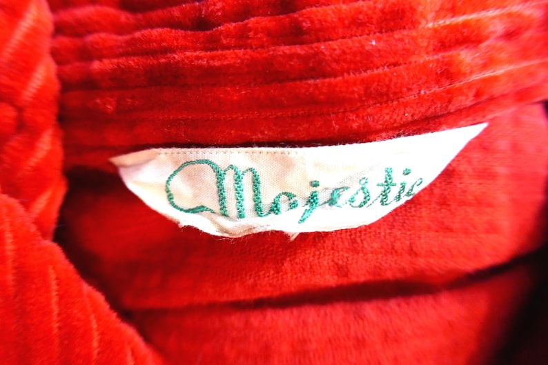 May include: A bright red corduroy garment featuring a white fabric label sewn into the collar area. The label displays the word Majestic embroidered in a cursive green font. The corduroy fabric has a distinct ribbed texture throughout the surface.
