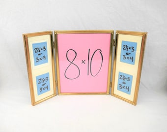 May include: A gold-toned metal triptych picture frame with three hinged panels. The center panel displays the text 8x10 in black marker on a pink background. The two side panels each contain two blue inserts with the text 2 1/2 x 3 or 3x4.