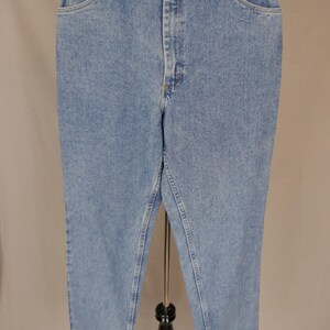 May include: Light blue denim jeans with a classic fit, featuring a button and zipper closure, front and back pockets, and a small logo on the front pocket. The jeans are displayed on a black stand against a neutral background.