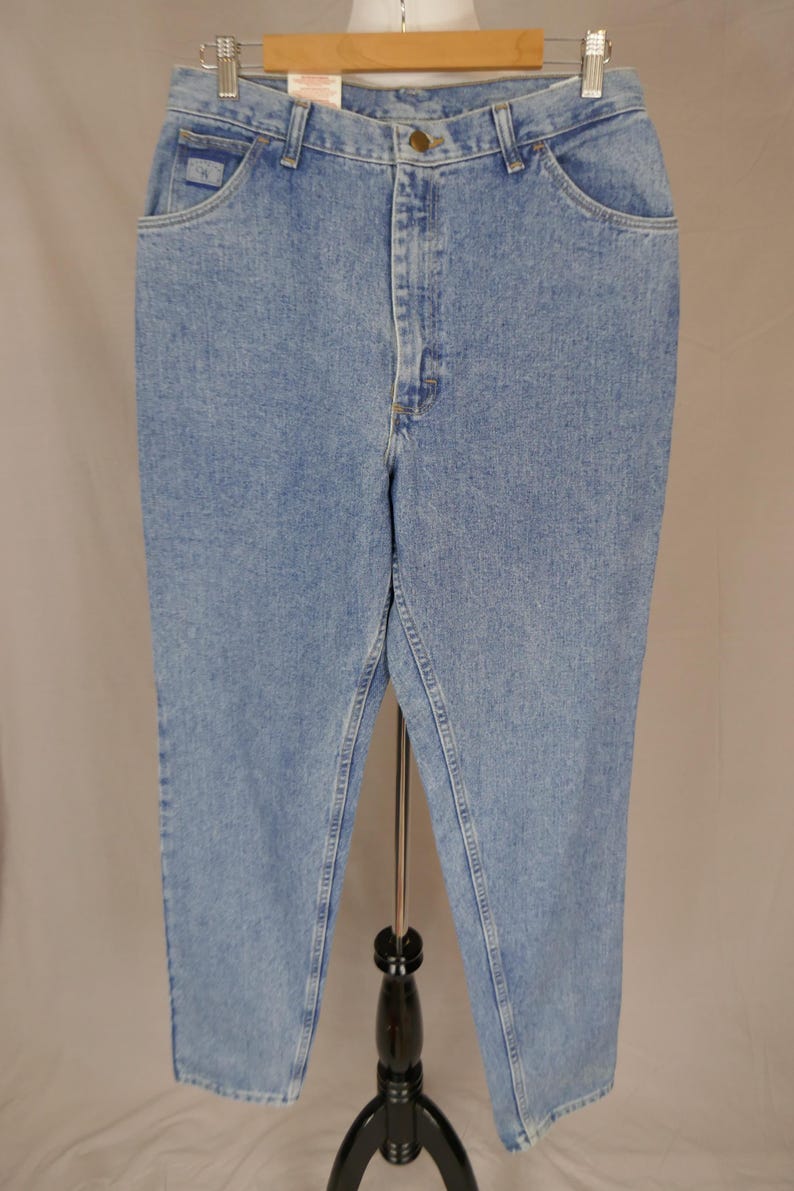 May include: Light blue denim jeans with a classic fit, featuring a button and zipper closure, front and back pockets, and a small logo on the front pocket. The jeans are displayed on a black stand against a neutral background.