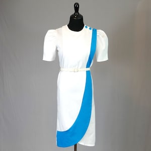 May include: White dress with short sleeves and a blue accent panel. The dress features a white belt and blue buttons on the shoulder. The dress is displayed on a black mannequin.