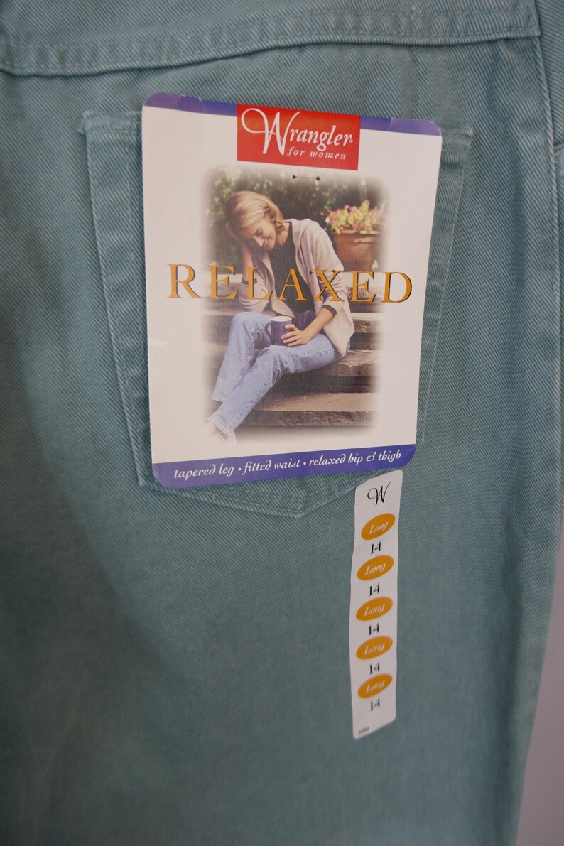 K&ouml;nnte beinhalten: Hellt&uuml;rkise Wrangler-Jeans mit einem Etikett mit der Aufschrift "Relaxed". Das Etikett zeigt ein Bild einer Person, die auf Stufen sitzt. Die Jeans hat ein schmales Bein, eine taillierte Taille und eine entspannte H&uuml;fte und Oberschenkel. Gr&ouml;&szlig;e 14.