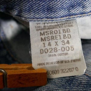 May include: Close-up of a denim jeans label with the text "14 X 34" and "100% Cotton". The label is attached to blue denim fabric. The jeans are held by a wooden hanger.