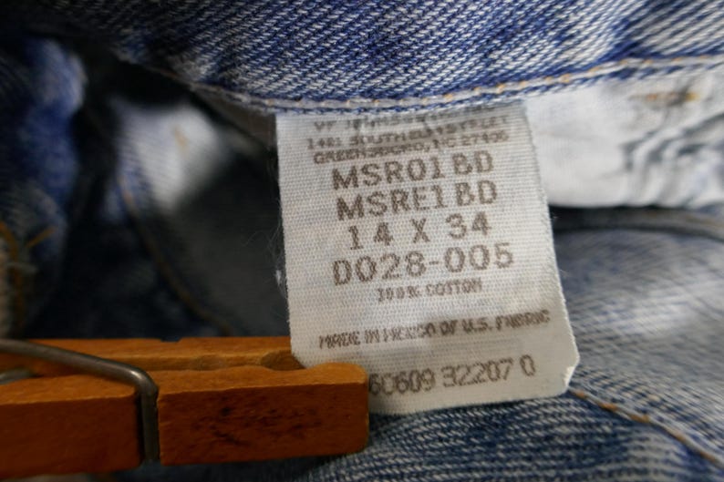 May include: Close-up of a denim jeans label with the text "14 X 34" and "100% Cotton". The label is attached to blue denim fabric. The jeans are held by a wooden hanger.