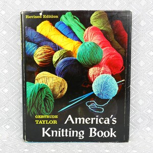 Peut inclure: Un livre relié intitulé America's Knitting Book de Gertrude Taylor, Revised Edition. La couverture noire présente un arrangement coloré d'écheveaux de laine jaunes, bleus, verts, rouges et orange, avec deux aiguilles à tricoter bleues posées dessus.