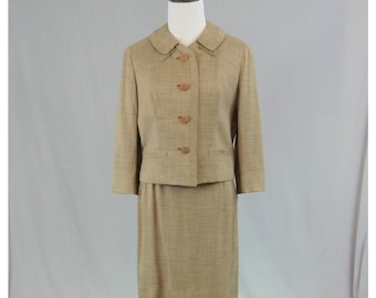 May include: A tan two-piece skirt suit featuring a cropped jacket with a rounded collar, three-quarter length sleeves, and four round brown buttons down the front. The matching skirt is knee-length with a straight cut and a subtle textured fabric finish.