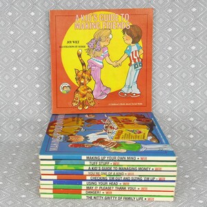 May include: A stack of children's books by Joy Wilt. The top book is titled A Kid's Guide to Making Friends, featuring an illustration of two children and a cat. The spines of the books below list titles including Making Up Your Own Mind, Tuff Stuff, A Kid's Guide to Managing Money, You're One of a Kind, Checking 'Em Out and Sizing 'Em Up, Using Your Head, May I? Please? Thank You!, Danger!, and The Nitty-Gritty of Family Life.