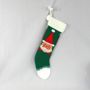 May include: A green knitted Christmas stocking featuring a central graphic of Santa Claus wearing a red hat. The stocking has a white textured cuff at the top, a white toe section, and a small white pom-pom attached to a white hanging loop.