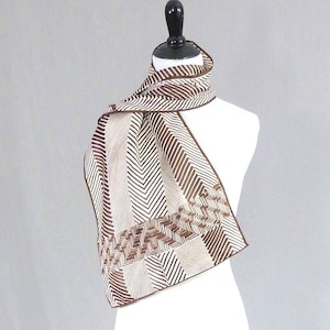 May include: A rectangular scarf featuring a brown and cream herringbone pattern with alternating vertical and horizontal sections. The scarf has a solid brown border along the edges and is draped around a white mannequin neck form.