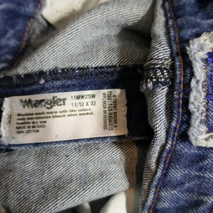 May include: Close-up of blue denim Wrangler jeans. The jeans have a label with the brand name and size 11/12 x 32. The jeans have a distressed look with visible stitching and a blue embroidered logo.