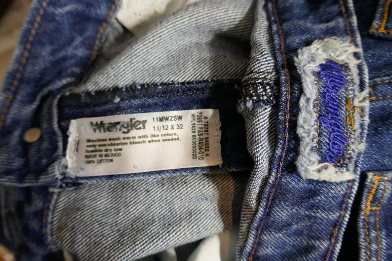 May include: Close-up of blue denim Wrangler jeans. The jeans have a label with the brand name and size 11/12 x 32. The jeans have a distressed look with visible stitching and a blue embroidered logo.