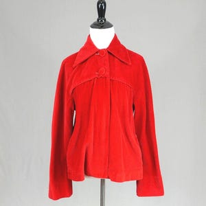 May include: A bright red corduroy jacket featuring a pointed collar, long sleeves, and a front button closure with two visible matching red buttons. The garment has a structured yoke detail across the chest and a straight hemline, displayed on a mannequin.