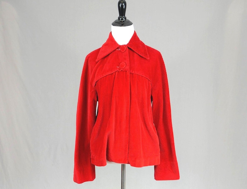 May include: A bright red corduroy jacket featuring a pointed collar, long sleeves, and a front button closure with two visible matching red buttons. The garment has a structured yoke detail across the chest and a straight hemline, displayed on a mannequin.