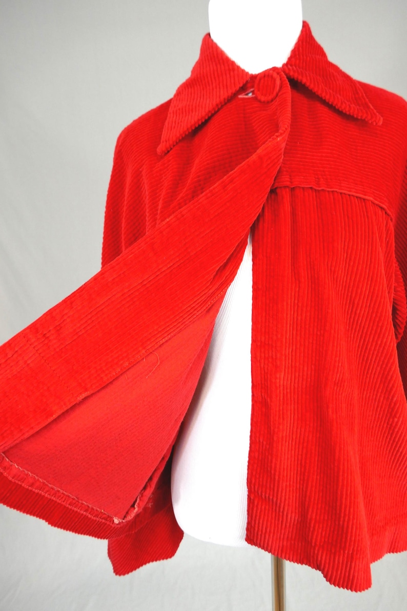 May include: A bright red corduroy jacket featuring a wide collar, a single large fabric-covered button closure at the neckline, and a flared silhouette. The jacket is made of a textured ribbed fabric and is displayed on a white mannequin form.