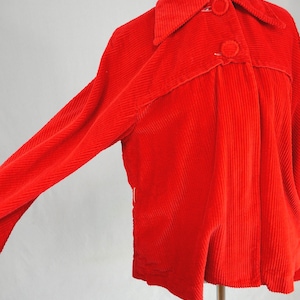 May include: A bright red corduroy jacket featuring a classic collar, long sleeves, and two large matching fabric-covered buttons on the front placket. The garment is made of a textured ribbed fabric and is displayed on a mannequin torso.