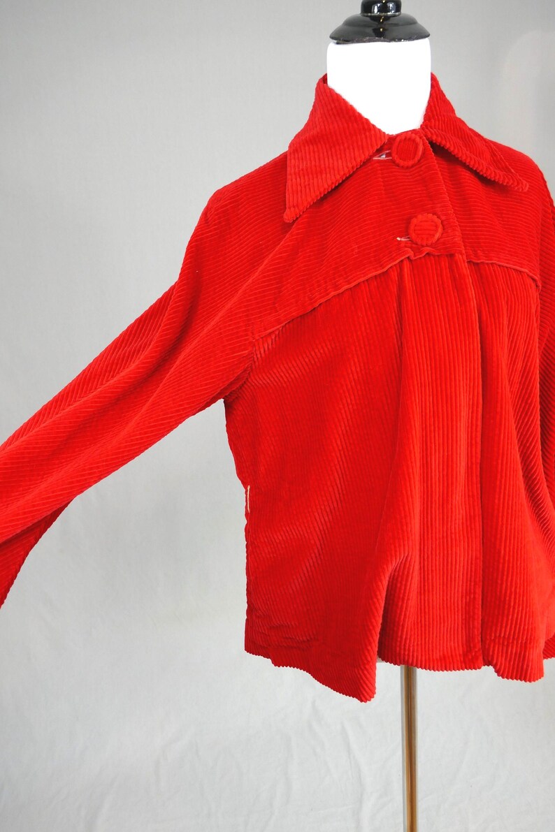 May include: A bright red corduroy jacket featuring a classic collar, long sleeves, and two large matching fabric-covered buttons on the front placket. The garment is made of a textured ribbed fabric and is displayed on a mannequin torso.