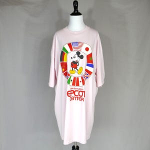 以下が含まれることがあります： 薄いピンク色の半袖Tシャツ。中央には様々な国旗が円状に配置され、その中にミッキーマウスのグラフィックが描かれています。グラフィックの下には、Walt Disney World EPCOT CENTERという文字が太い赤色のフォントでプリントされています。