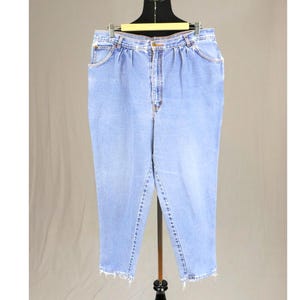 Peut inclure: Jean en denim bleu clair avec une taille haute, des plis sur le devant, une fermeture à bouton et à glissière, et un ourlet effiloché au bas des jambes. Le denim présente un aspect délavé avec des coutures visibles sur l'ensemble.