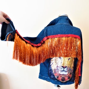 May include: A blue denim jacket with a red sequined trim and orange fringe. A lion graphic is on the back of the jacket.