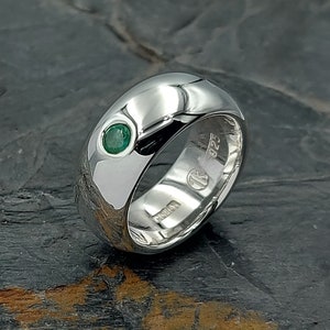May include: A polished silver band ring featuring a single, small, round, green gemstone set flush into the metal. The interior of the band is clearly stamped with the hallmark 925, indicating sterling silver, along with other maker marks.