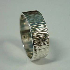 Pode incluir: Um anel de banda de prata com uma superfície texturizada e estriada. O anel é largo e tem um acabamento polido.