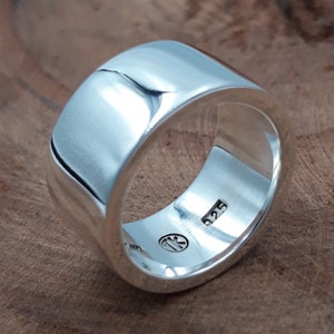 May include: A wide, polished silver band ring with a smooth, reflective surface. The interior of the band features a stamped 925 hallmark indicating sterling silver content and a circular maker's mark with the letters TK inside the metal band.