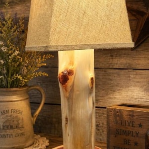May include: A table lamp featuring a natural light-colored wood log base resting on a circular wood slice stand. The lamp is topped with a square, textured beige burlap lampshade that emits a warm, soft glow from the light source inside.