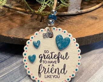 May include: A white, scalloped-edge ornament with the text "So grateful to have a friend like you." It features three blue heart accents and a small silver charm with "2021." The ornament is suspended by a twine string with silver and blue beads.