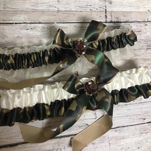 May include: Two elastic garters featuring a camouflage print ribbon and a solid white or sheer white ruffled trim. Each garter is adorned with a camouflage ribbon bow at the center, which is accented by a small, metallic brown flower embellishment.