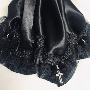 May include: A black satin shawl with a pleated lace trim and a decorative cross pendant. The shawl has a soft, flowing texture and is embellished with black lace and small pearl accents. The cross pendant is encrusted with sparkling crystals.