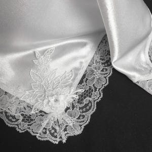 May include: Close-up of a white satin fabric with delicate lace trim. A floral applique with a small gem is attached to the fabric. The lace and applique add a touch of elegance to the textile.