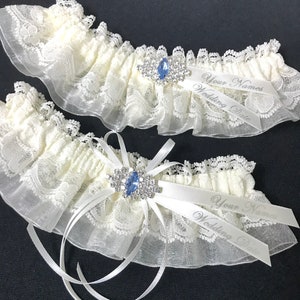 May include: Two ivory lace garters with blue rhinestone embellishments and satin ribbons. The ribbons are personalized with the text "Your Names Wedding Date".