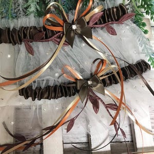May include: Two elastic garters featuring dark brown satin bands and white tulle ruffles adorned with small clear rhinestones. Each garter is decorated with a central dark brown satin bow, orange and gold ribbons, and small brown fabric leaves.