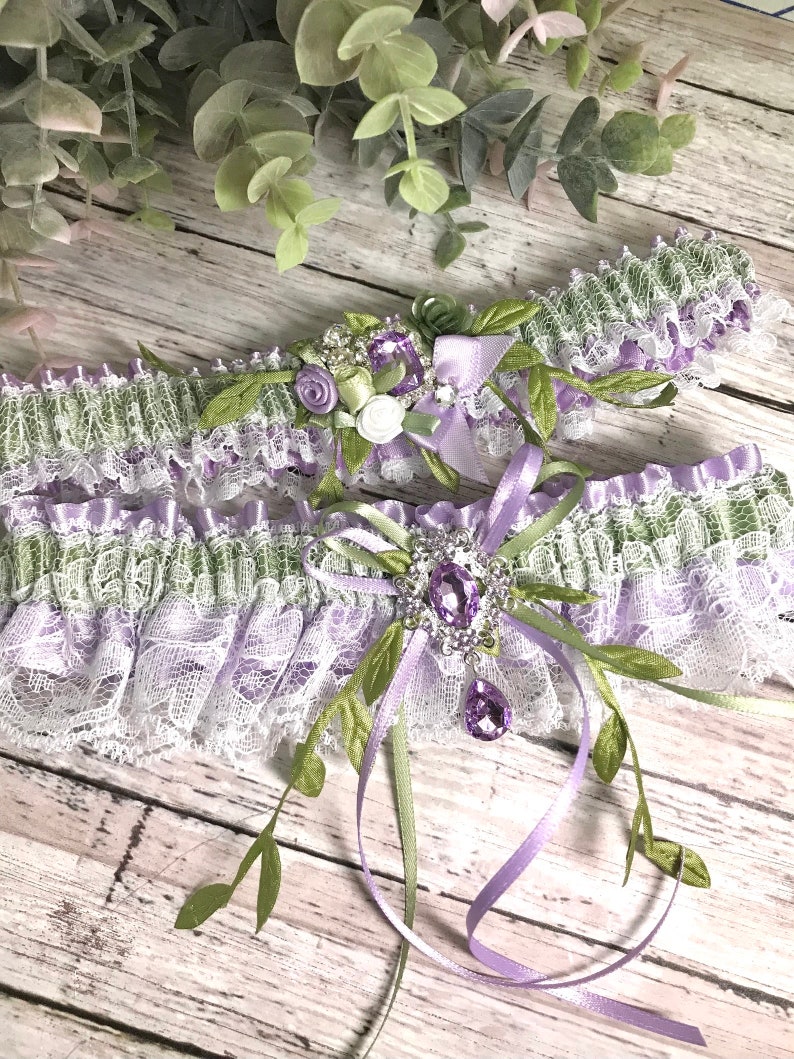 May include: A delicate white and lavender lace garter with green satin ribbon and a crystal embellishment. The garter has a floral design with white and green flowers.