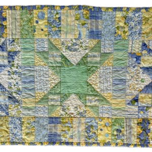 May include: A quilted table runner with a patchwork design. The runner features a central star pattern in shades of blue and green, surrounded by squares of yellow, blue, and white floral fabrics. The edges are trimmed with a yellow border.