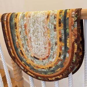 May include: A half-circle braided rug with a fall theme. The rug features a variety of colors, including brown, orange, yellow, and green. The design includes sunflowers and other fall foliage. The rug is draped over a wooden railing.
