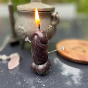 May include: A dark purple candle with a flame burning at the top. The candle is decorated with several pins. The candle is sitting on a dark gray surface.