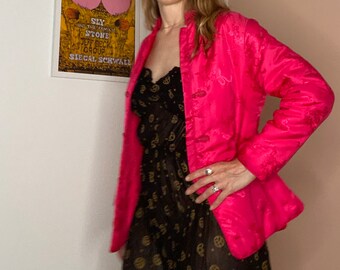 May include: A bright pink, long-sleeved jacket with a mandarin collar and decorative frog closures. The jacket has an embroidered floral pattern and is worn over a black dress with a gold pattern. A framed poster is visible in the background.