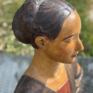 Puede incluir: Un busto de madera de una mujer con la cara y el pelo pintados. El busto está pintado en tonos marrones, rojos y dorados. El pelo de la mujer está recogido en un moño y lleva una prenda marrón y roja.