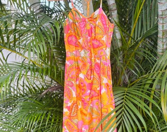 May include: A colorful, patterned dress with a halter neckline and spaghetti straps hangs on a wooden hanger. The dress features a vibrant paisley design in shades of pink, orange, and yellow. The dress is approximately 40 inches long.
