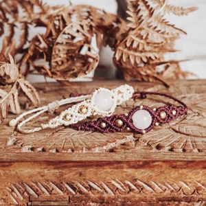 May include: Two bracelets with rose quartz stones and gold beads on a wooden box. The bracelets are made with macrame and have a white and burgundy color scheme.