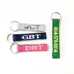 May include: Four keychains with different colored fabric straps and embroidered text. The first keychain has a gray strap with the text "JCT" in rainbow colors. The second keychain has a navy blue strap with the text "GBT" in white. The third keychain has a pink strap with the text "DRT" in white. The fourth keychain has a green strap with the text "864578-8614" in yellow.