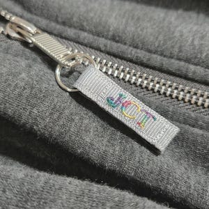 May include: A close-up of a zipper pull with a gray fabric tag that says "J.C.I.T." in rainbow colored thread.