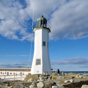 May include: A white lighthouse with a green roof stands on a rocky shore. The lighthouse is surrounded by small piles of stones, and the sky is blue with white clouds.