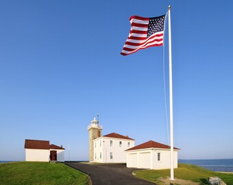 May include: A white lighthouse with a red roof and a tall white flagpole with an American flag waving in the wind. The lighthouse is on a grassy hill with a white building with a red roof in the foreground.