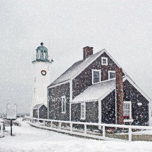 May include: A white lighthouse with a green roof stands tall against a snowy backdrop. A dark gray wooden house with white trim sits beside the lighthouse. Snow falls softly, creating a winter wonderland scene.