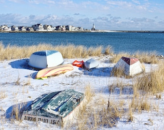 May include: Five small boats on a snowy beach with a view of a town and a lighthouse in the distance. The boats are various colors, including red, white, and blue. The beach is covered in snow and dry grass.