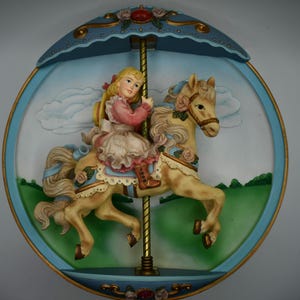Puede incluir: Un adorno de pared con temática de carrusel que representa a una niña montada en un caballo de color beige. El caballo tiene detalles florales y está ambientado sobre un fondo de cielo azul con nubes y hierba verde. La pieza está enmarcada en un óvalo azul y dorado.