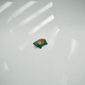 May include: A small, decorative ceramic frog figurine. The frog is primarily teal with orange and red accents. It has black feet and is positioned on a white surface.